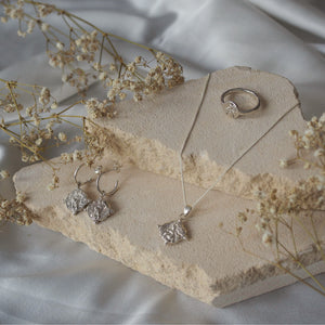 A silver jewelry set, is displayed on a beige stone with delicate white flowers surrounding it.