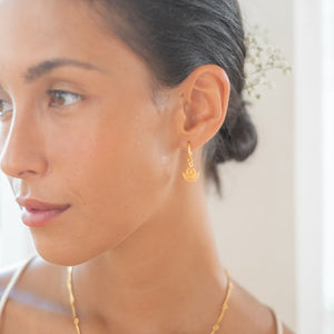 A close-up of a woman with gold sunburst earrings and a gold necklace, looking off to the side.