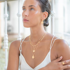 A woman looking up wearing a gold hoop earring and a multiple gold necklaces,  with her hair styled in a low bun adorned with small white flowers.