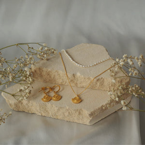 A jewelry display featuring gold earrings, a gold necklace with a circular sunburst pendant, and a pearl necklace arranged on beige stone pieces with dried flowers.