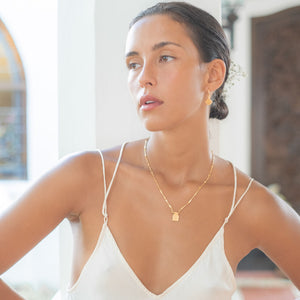 A woman with brown hair is wearing a gold necklace and golden earrings with a white dress