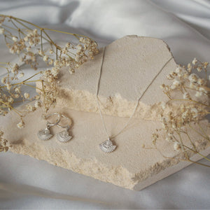 A jewelry display featuring silver earrings, a gold necklace with a circular sunburst pendant, arranged on beige stone pieces with dried flowers.