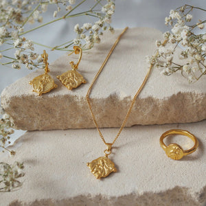 A gold jewelry set, including a pair of hoop earrings with religious figure design charms, a necklace with a matching pendant, and a ring with a similar design, is displayed on a beige stone with delicate  white flowers surrounding it.