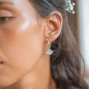 A close-up of a woman with silver sunburst earrings and a gold necklace, looking off to the side.