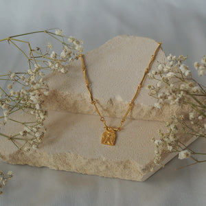 A gold necklace, displayed on a light stone surface with some dried plants in the background.