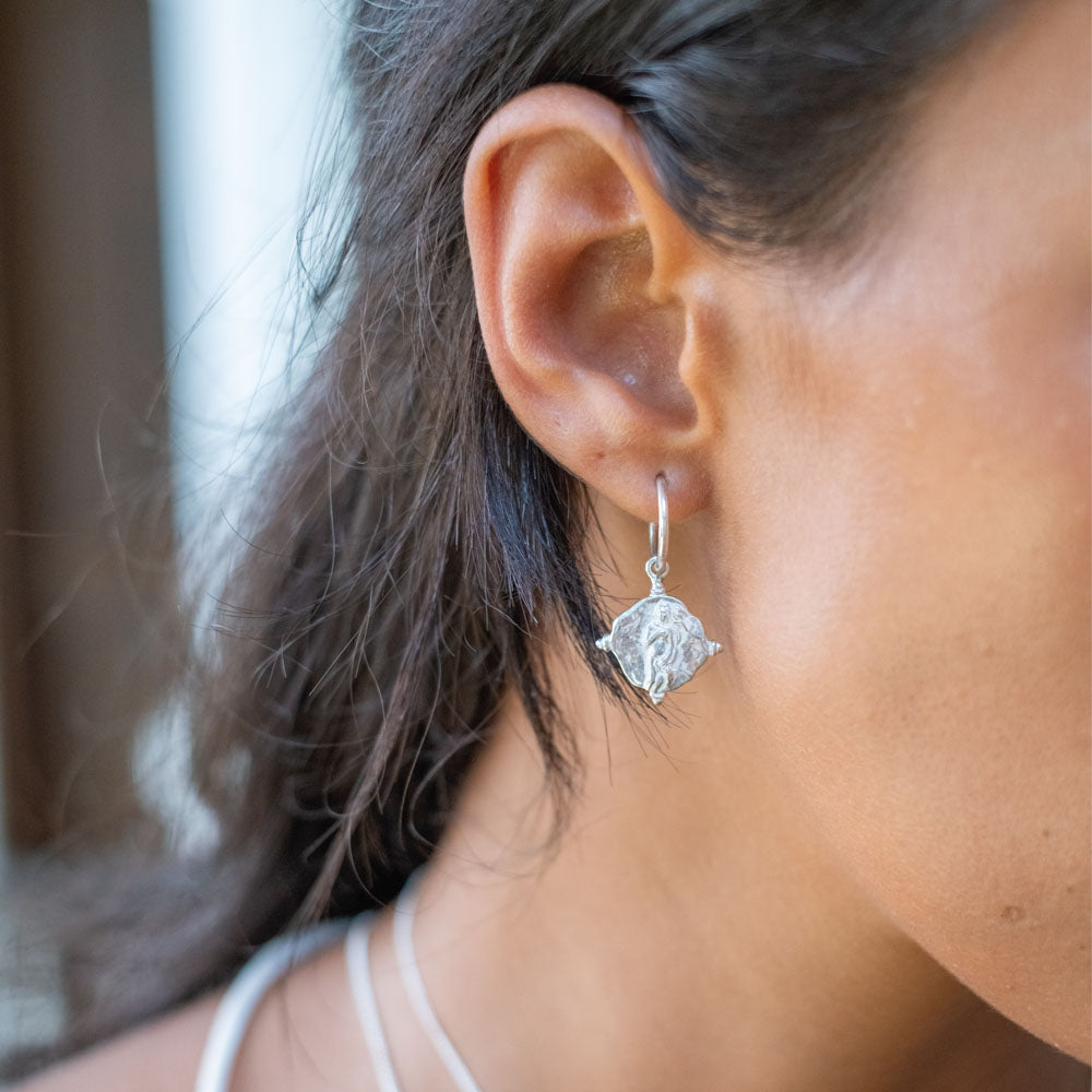 A pair of silver hoop earrings featuring religious figure designs on hanging charms.