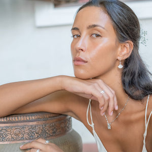 A woman with black hair wearing silver earrings, necklace and rings while holding her arm on an object