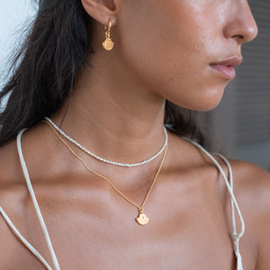  A close-up of a woman wearing gold sunburst earrings, a gold necklace with a sunburst pendant, and a pearl necklace.