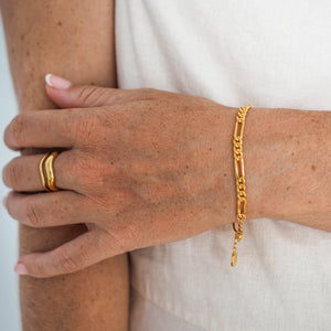 A person wearing a gold link chain bracelet and a gold ring featuring two stacked designs on their fingers.