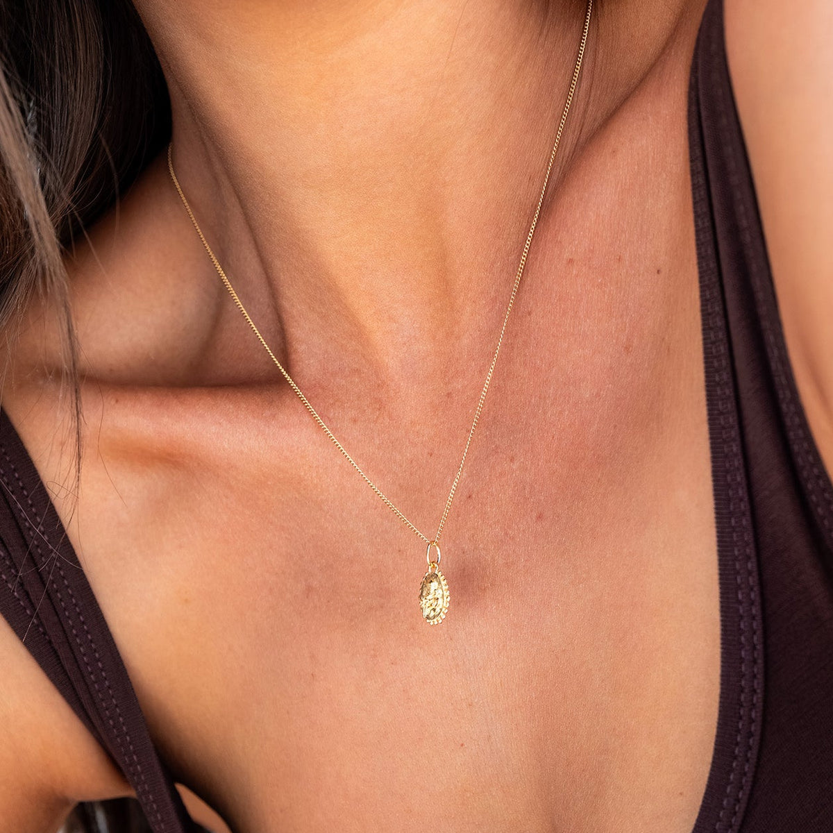 A close-up lifestyle shot of a model wearing a delicate gold chain necklace with a small oval pendant, paired with a dark brown tank top.