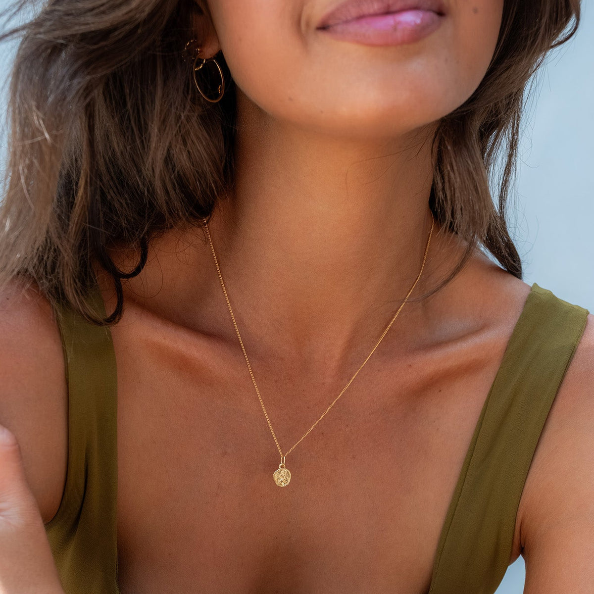 A close-up lifestyle shot of a woman wearing a delicate gold chain necklace with a small round disc pendant, styled with an olive green top against a light blue background.