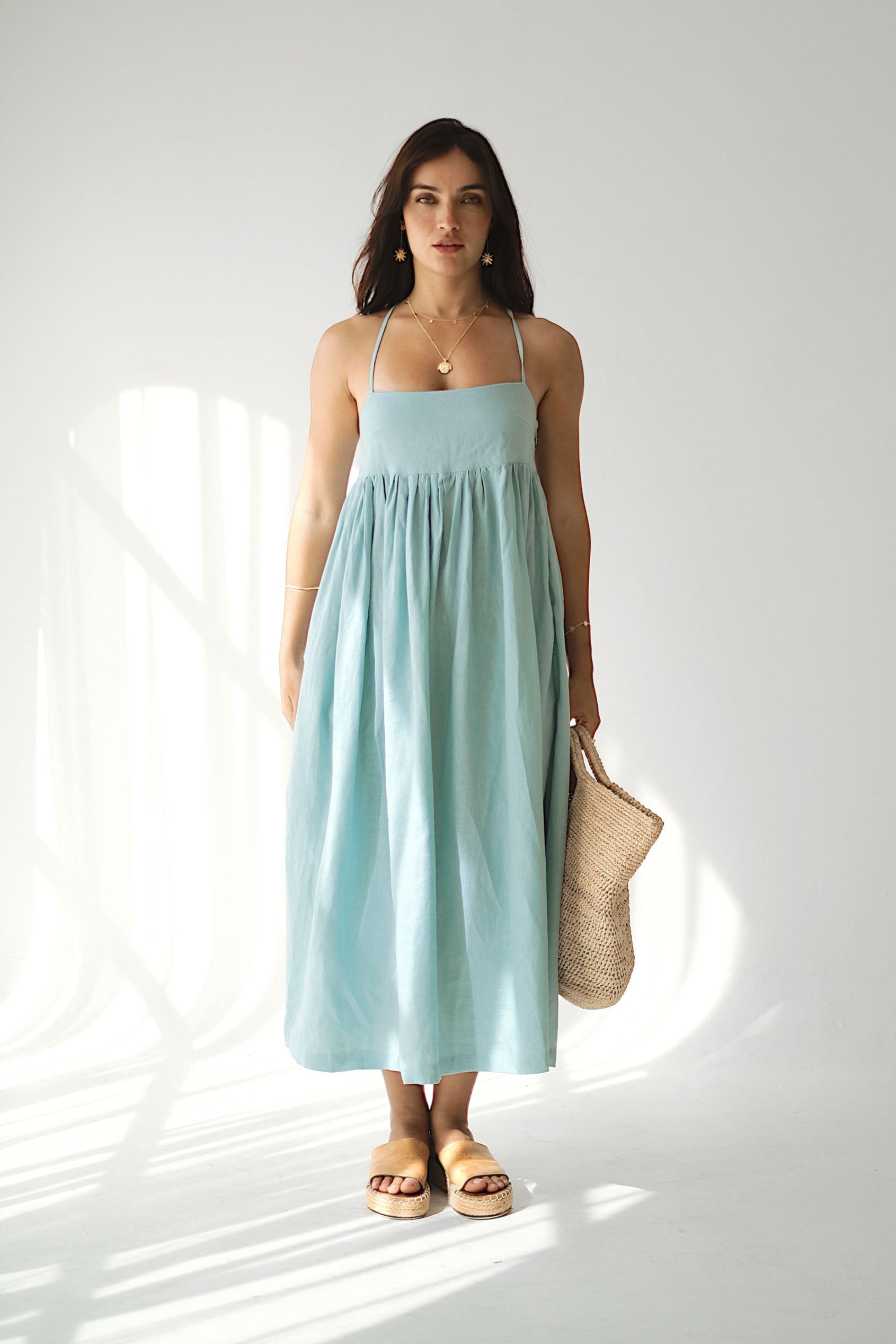 Woman wearing a light blue dress holding a beige bag in a bright room.