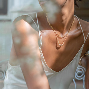 A person wearing a white tank top and two layered gold necklaces, one featuring a crescent moon pendant and the other with elongated chain links, captured in soft sunlight.