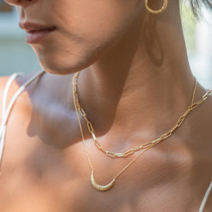 A close-up of a person wearing two layered gold necklaces, one with a crescent moon pendant and the other with elongated chain links, along with a small gold hoop earring.