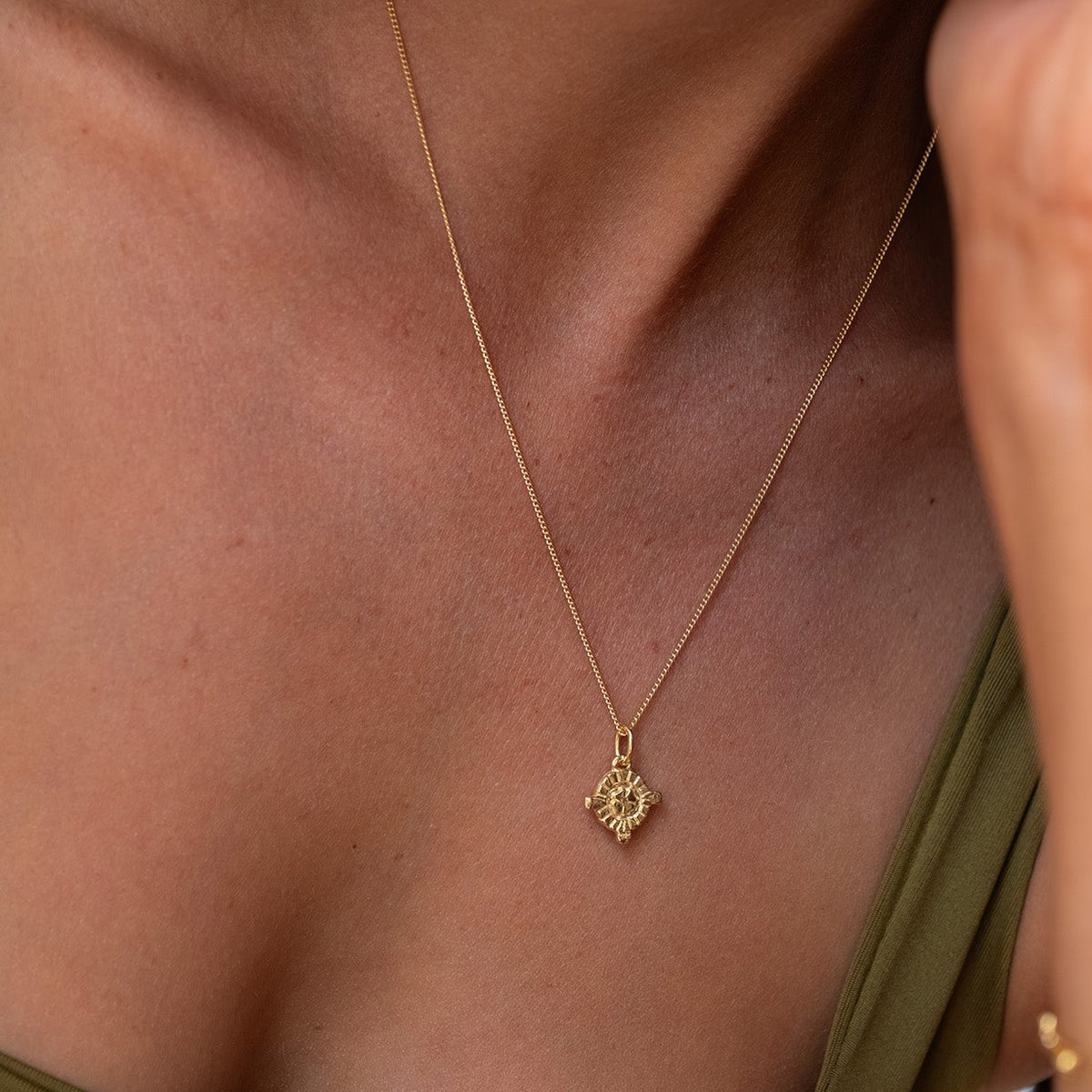 A close-up lifestyle shot of a woman wearing a delicate gold necklace with a small Hamsa hand pendant, styled with an olive green top.