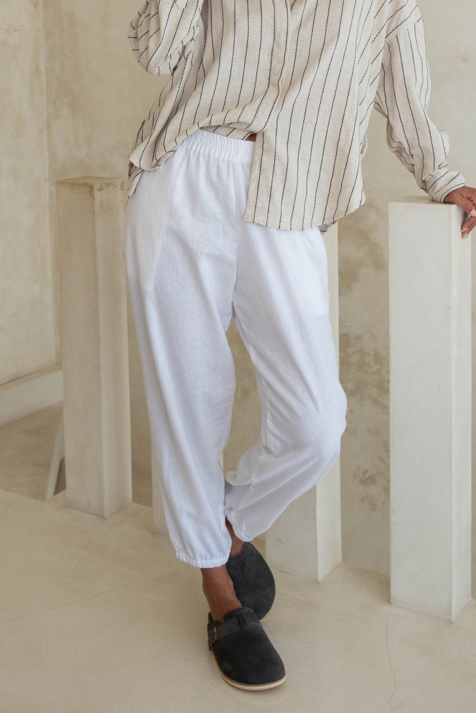 A person leaning against a railing, wearing a striped shirt and white pants with black slip-on shoes.