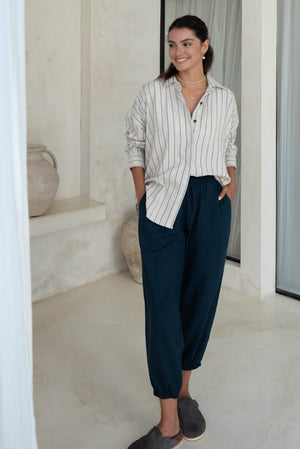 A woman stands wearing a striped shirt and dark blue loose pants with blue slip-on shoes, accessorized with gold jewelry.