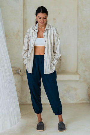 A woman wears a striped shirt over a white crop top and dark blue loose pants with blue slip-on shoes.