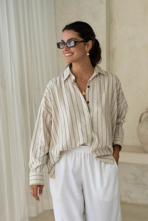 A person smiling, touching sunglasses, wearing a striped shirt and white pants.