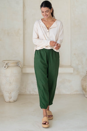 A woman in a beige long-sleeve button-up top and green loose pants with tan wedge sandals smiles while looking down.