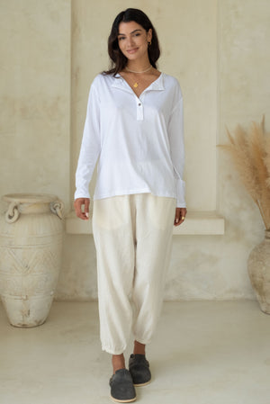 A woman stands wearing a white long-sleeve button-up top and light-colored loose pants with black slip-on shoes, accessorized with gold jewelry.