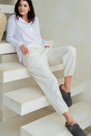 A woman is seated on white stairs, dressed in a white long-sleeve button-up top, light-colored loose pants, and blue slip-on shoes.