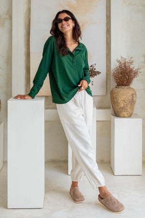 A woman wears a dark green long-sleeve top with white loose pants and beige slip-on shoes, accessorized with sunglasses and necklaces.