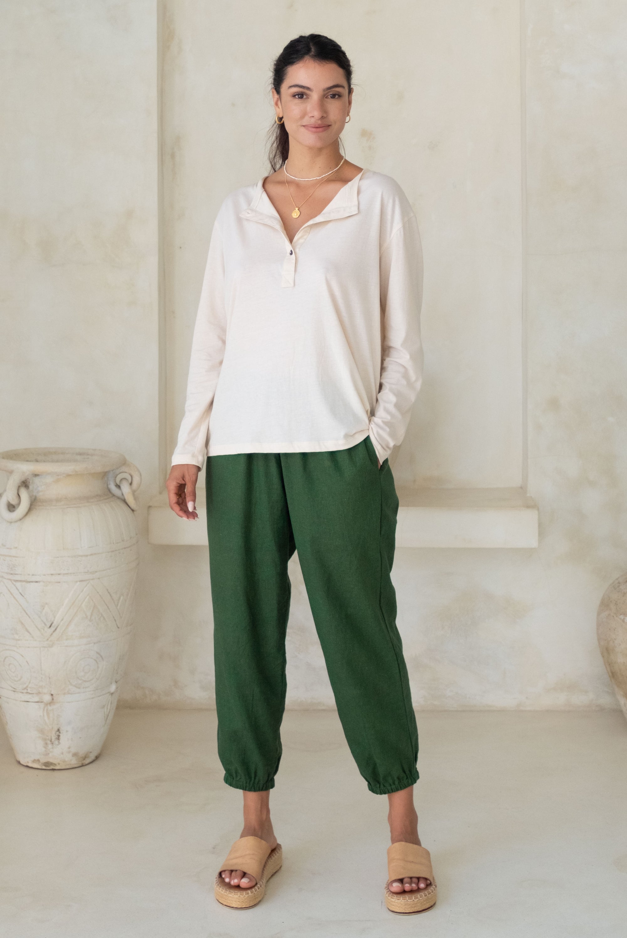 A woman stands wearing a beige long-sleeve button-up top and green loose pants with tan wedge sandals.