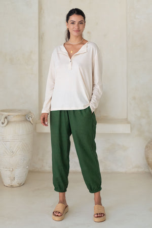 A woman stands wearing a beige long-sleeve button-up top and green loose pants with tan wedge sandals.