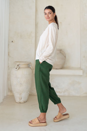 A side view of a woman wearing a beige long-sleeve button-up top with green loose pants and tan wedge sandals.