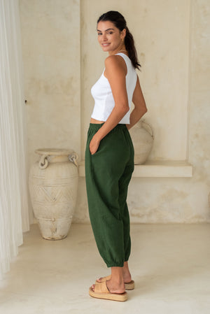 A woman wearing a white sleeveless crop top and green loose pants with tan wedge sandals.