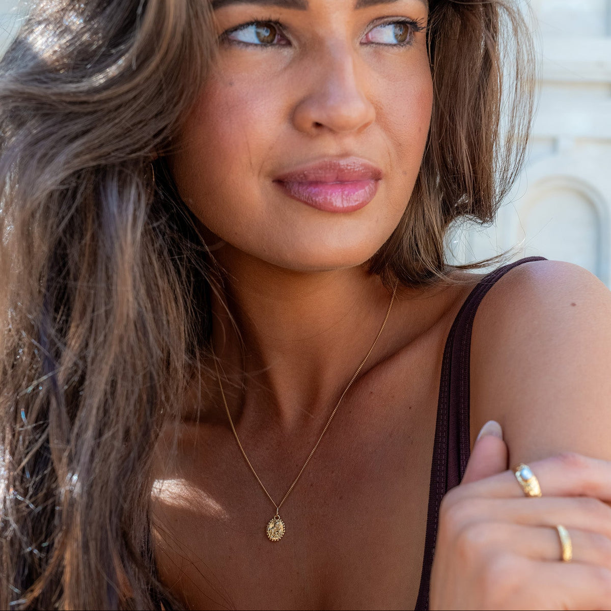 A close-up lifestyle shot of a woman with long brown hair wearing a delicate gold chain necklace with a small round disc pendant, along with gold rings on her fingers. She is wearing a dark plum-colored top against a softly blurred light-colored background.