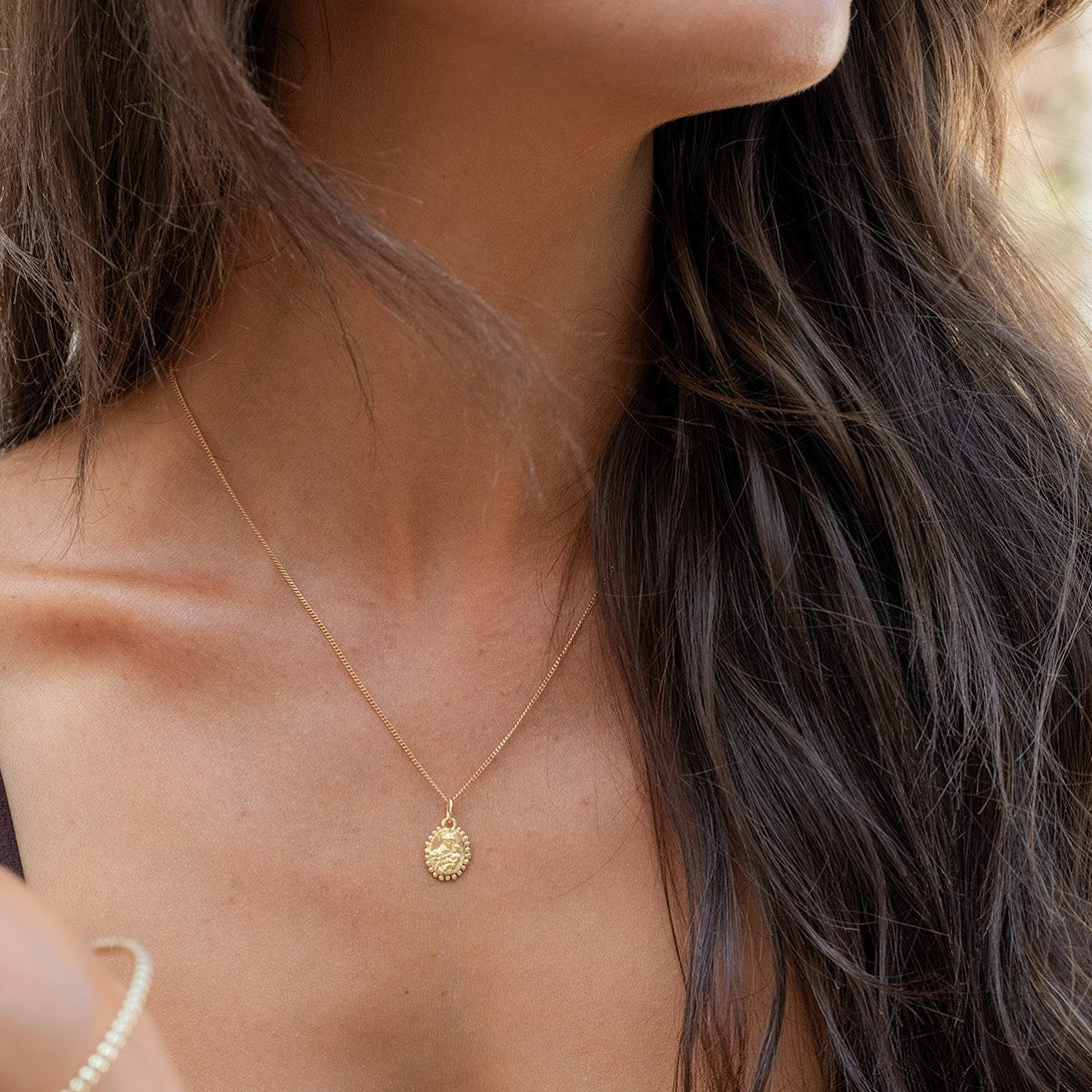 A close-up lifestyle shot of a woman wearing a delicate gold chain necklace with a small round disc pendant, styled with a gold bracelet and rings, wearing a dark brown ribbed tank top.