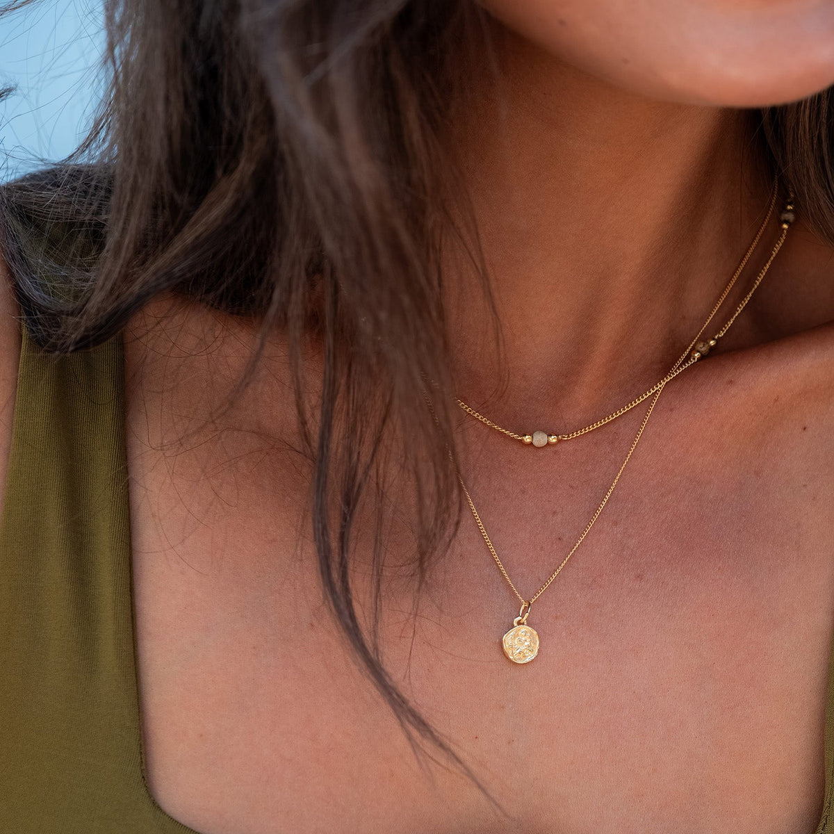 A close-up lifestyle shot of a model wearing a delicate gold double-layer necklace with a small round disc pendant, paired with a green top.