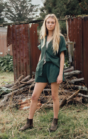 A woman in an oversized green button-up shirt and matching green shorts, along with brown ankle boots, stands in a grassy area with a rusty metal backdrop and woodpile.