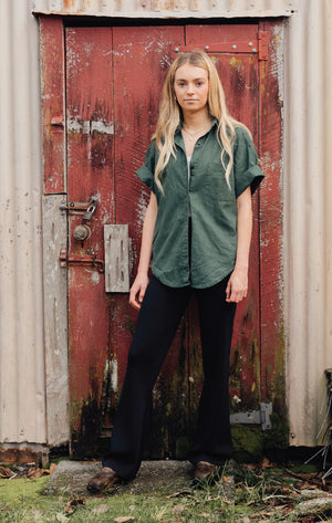 A woman in an oversized green button-up shirt, black wide-leg pants, and brown ankle boots, stands in front of a red rusted door.