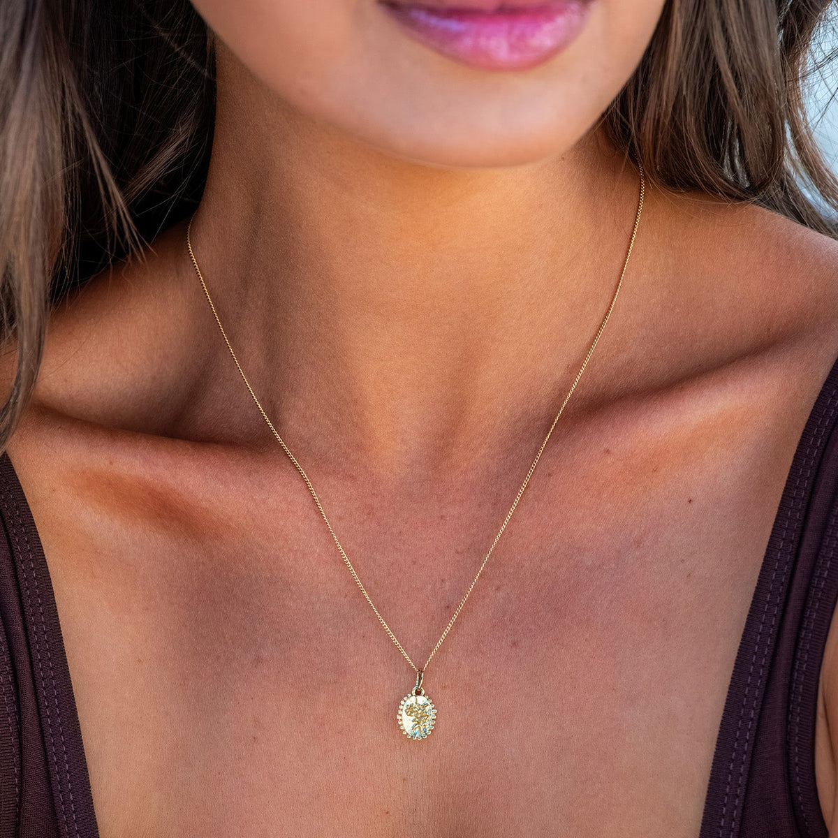 A close-up lifestyle shot of a woman wearing a delicate gold chain necklace with a small oval pendant featuring a green stone, paired with a dark brown scoop-neck top.
