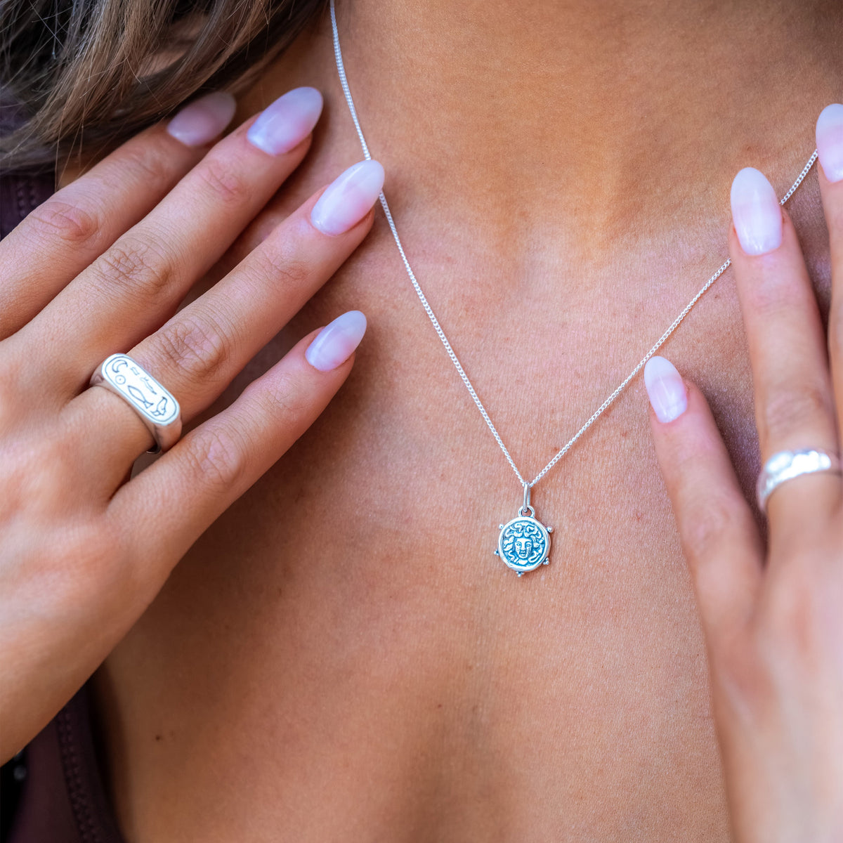 A close-up lifestyle shot of a woman wearing a delicate silver chain necklace with a round, light blue stone pendant. Her hands are gently framing the necklace, and she is wearing a dark plum-colored top.