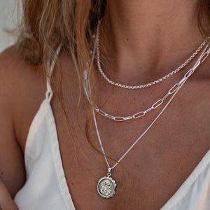 Close-up of layered silver necklaces with intricate pendants on a woman's neck, who is wearing a white strappy top.