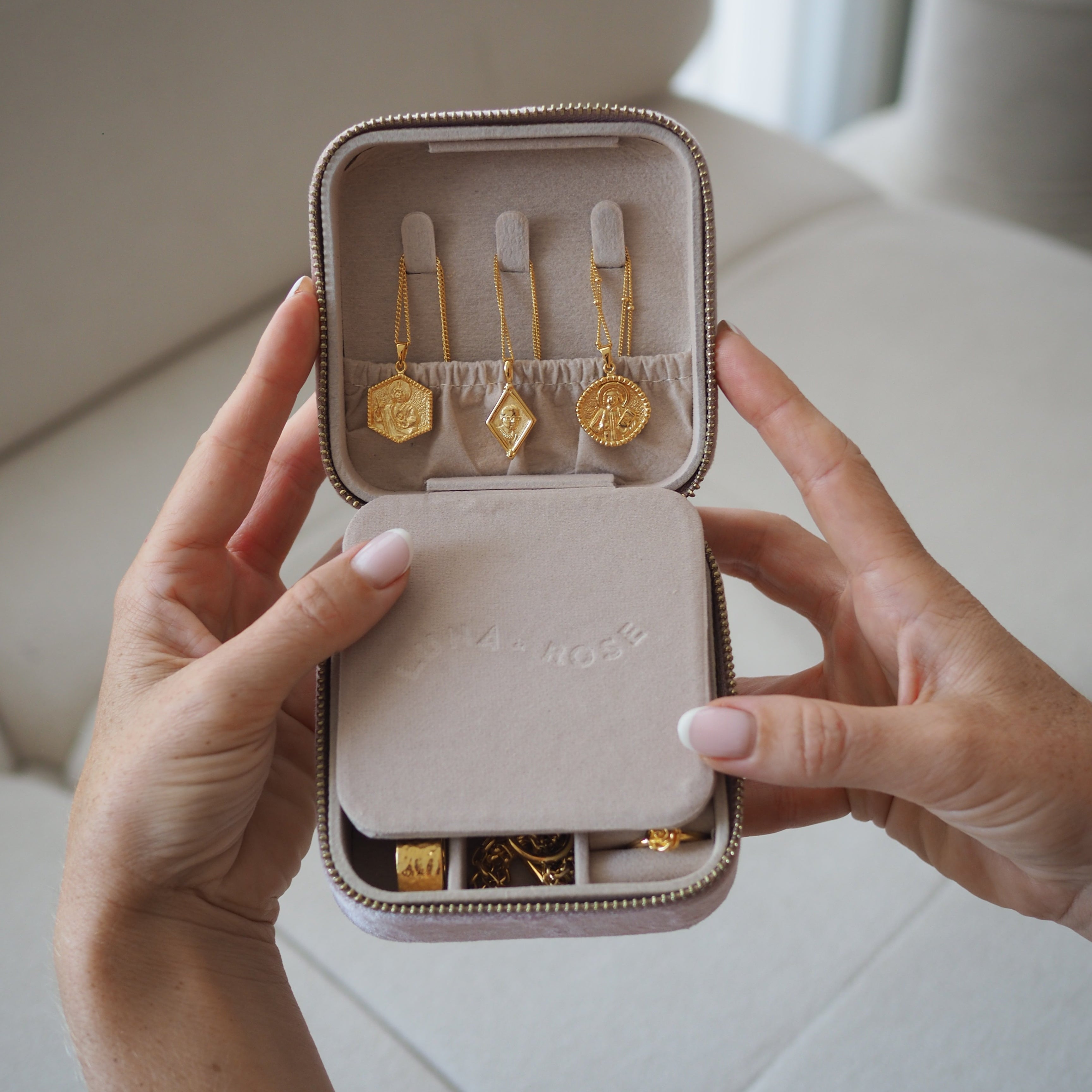 A person holds open a small zippered jewelry case with gold earrings displayed inside. The case is a soft blush pink velvet with a silver zipper, and the background is a neutral light beige sofa.