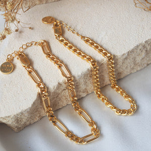 Two gold chain bracelets with clasps, laid on textured stones, featuring engraved tags.