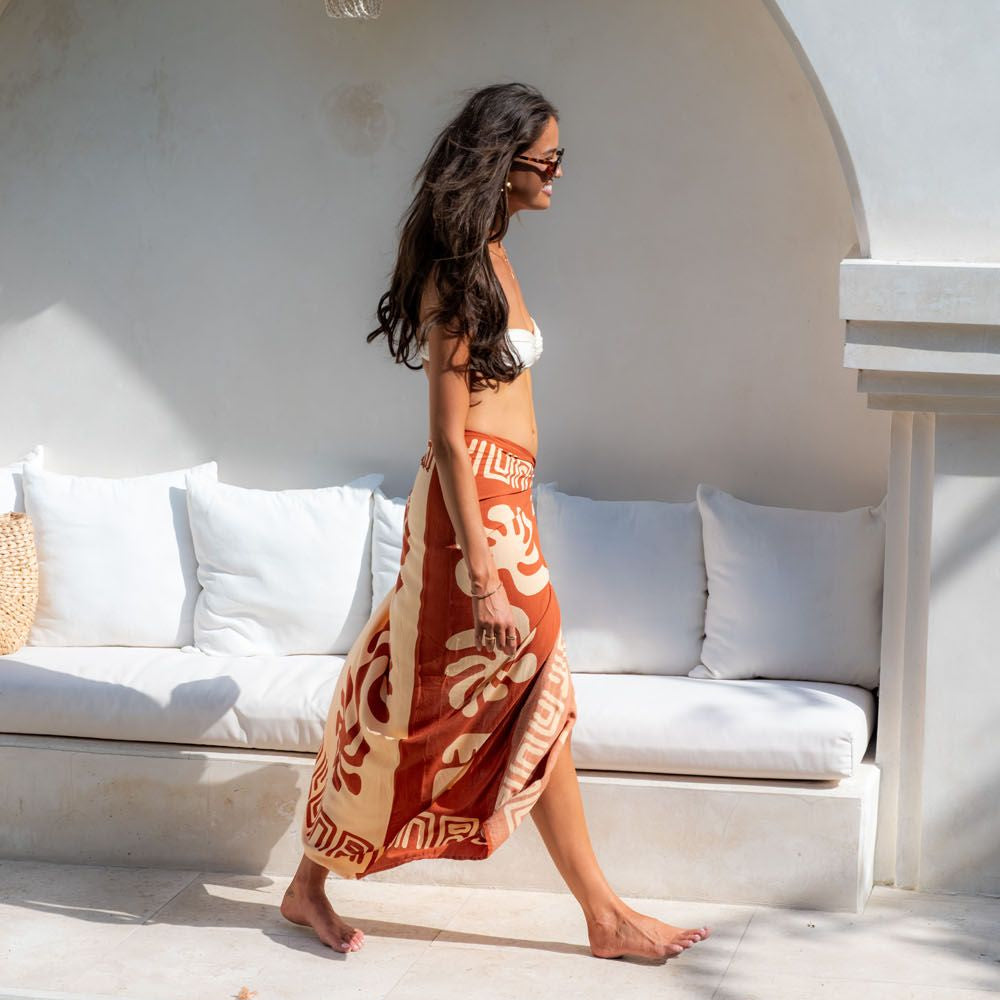 A woman walks barefoot beside a swimming pool in front of a white arched alcove with a cushioned bench. She is wearing a white bikini and a long rust-orange and cream patterned wrap skirt or sarong.