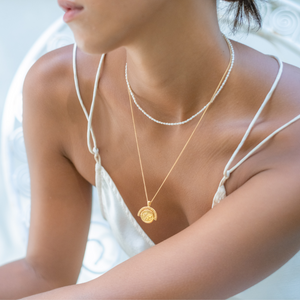 A close-up of a woman wearing a layered pearl and gold pendant necklace over a white satin top with spaghetti straps.