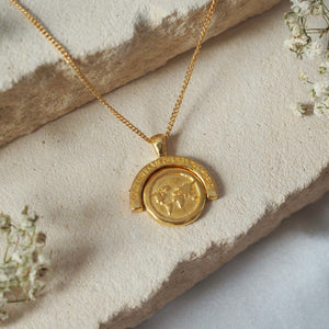 A gold necklace with a world map pendant and the inscription "OUR LOVE CAN CHANGE THE WORLD" on a stone background.