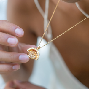 A close-up of a woman holding a gold necklace with a pendant that reads "OUR LOVE CAN CHANGE THE WORLD" while wearing a white dress.