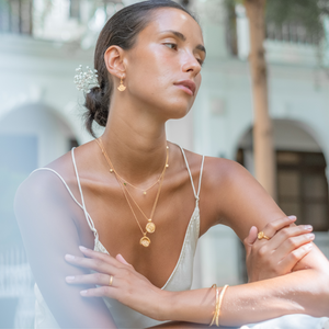 A woman wearing layered gold necklaces with matching earrings, rings, and a bracelet, dressed in a white top and seated in an elegant setting.