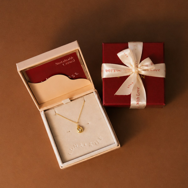 A gold necklace with a small round pendant displayed in an open cream-colored jewelry box, alongside a red gift box tied with a cream ribbon that reads 'With Love'.