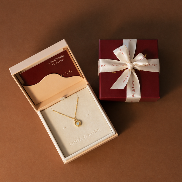 A gold necklace with a circular pendant displayed inside an open cream jewelry box, alongside a wrapped dark red gift box tied with a white ribbon on a brown background.