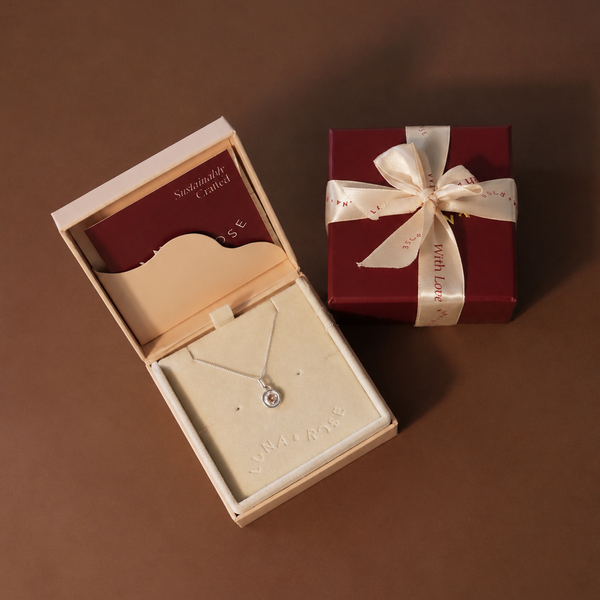 A luxury jewelry gift set featuring an open cream-colored box displaying a delicate silver necklace on a cream velvet insert, alongside a dark red square gift box tied with a cream satin ribbon.