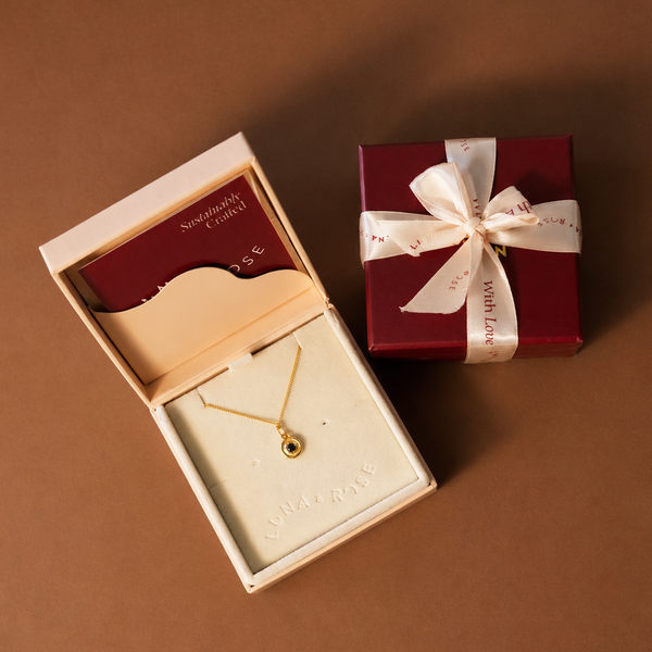 A gold necklace with a small round bead pendant displayed in an open cream jewelry box, alongside a dark red gift box tied with a white ribbon printed with With Love